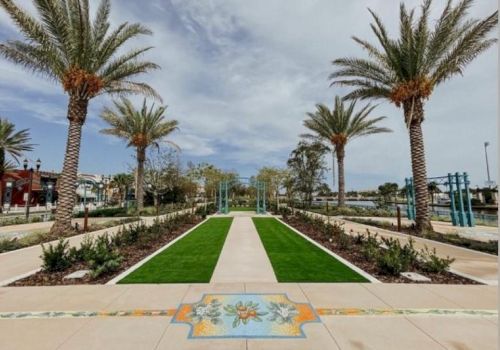 A garden walkway flanked by tall palm trees leads to a green artificial-turf play area with colorful tiles.