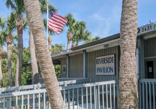 A Riverside Pavilion sign on a building behind palm trees, with an American flag flying above it.
