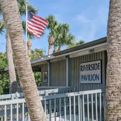 A Riverside Pavilion sign on a building behind palm trees, with an American flag flying above it.