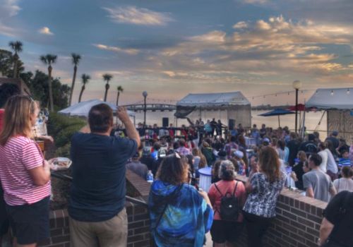 A crowd enjoys an outdoor event or concert by the water under a vibrant sky, with tents, palm trees, and a bridge in the background.