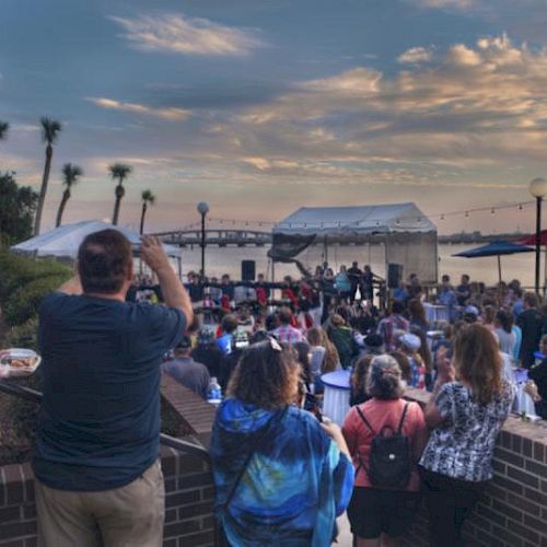 A crowd enjoys an outdoor event or concert by the water under a vibrant sky, with tents, palm trees, and a bridge in the background.