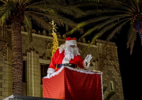 A man dressed as Santa in red waves while standing at a lit outdoor podium beside palm trees and festive lights.