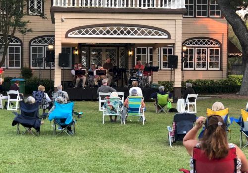 A small outdoor concert with people seated on lawn chairs facing a stage set up in front of a large house.