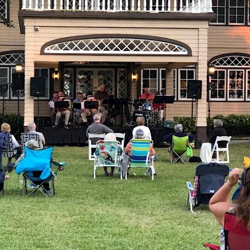 A small outdoor concert with people seated on lawn chairs facing a stage set up in front of a large house.