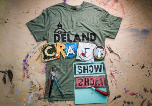 A T-shirt with "DELAND," colorful letters spelling "CRAFT," tools, and "SHOW" signs are on a messy, paint-stained surface.