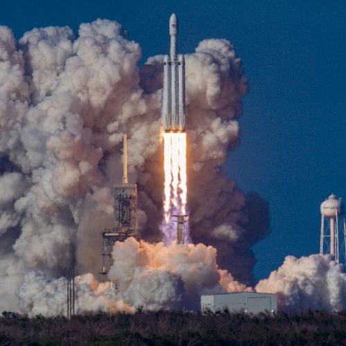 A rocket is launching into space amid large clouds of smoke and fire from the engines, with clear blue skies and structures visible in the background.