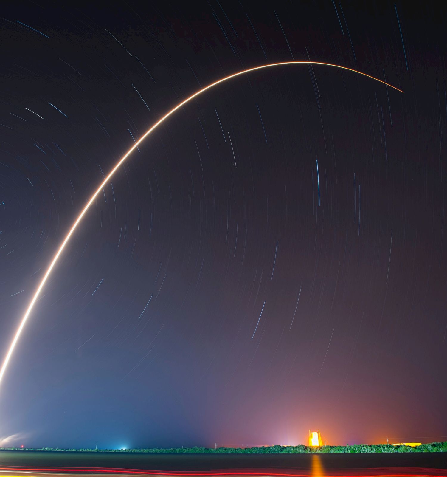 A rocket launch captured in long exposure, with a bright curved light arc and stars trailing in the night sky, displaying a vibrant landscape.