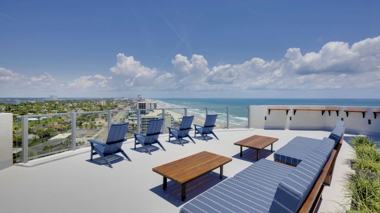 A rooftop terrace with blue lounge chairs and tables offers stunning ocean views under a bright, partly cloudy sky.