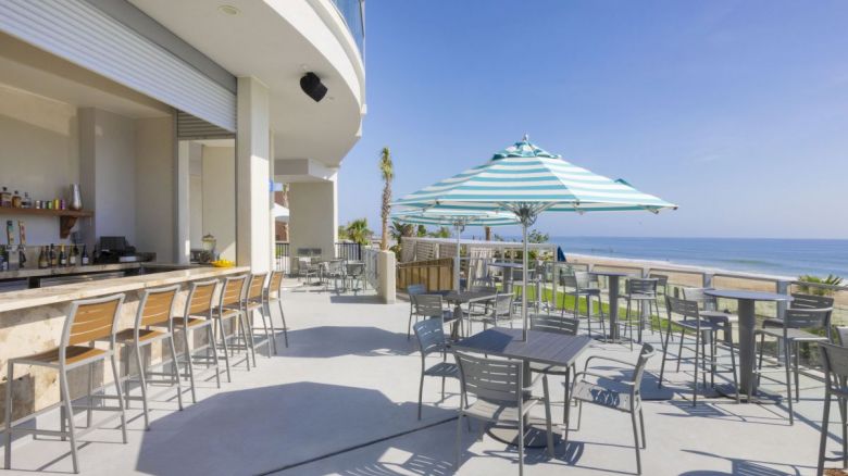 An outdoor bar and dining area next to a beach with tables, chairs, and an umbrella, offering a scenic ocean view in a sunny setting.