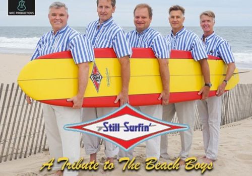 Five men in striped shirts hold surfboards on a beach. Text reads: "Still Surfin' - A Tribute to The Beach Boys."