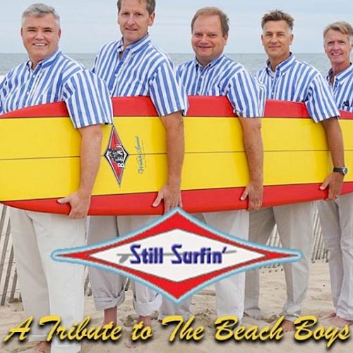 Five men in striped shirts hold surfboards on a beach. Text reads: "Still Surfin' - A Tribute to The Beach Boys."