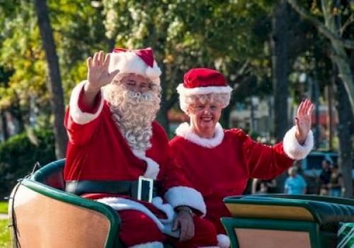 Two people dressed as Santa and Mrs. Claus are sitting in a sleigh, waving, with trees and people in the background.