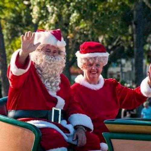 Two people dressed as Santa and Mrs. Claus are sitting in a sleigh, waving, with trees and people in the background.