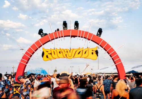 A crowded outdoor music festival with a huge red stage arch and spotlights, plus a colorful &ldquo;R&hellip;D&hellip;&rdquo; logo banner.