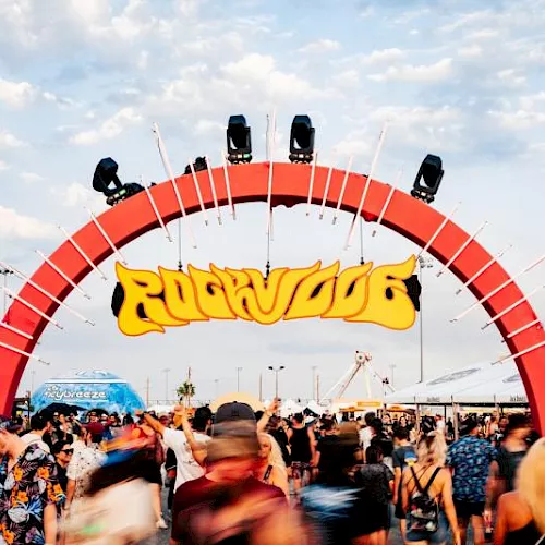 A crowded outdoor music festival with a huge red stage arch and spotlights, plus a colorful &ldquo;R&hellip;D&hellip;&rdquo; logo banner.