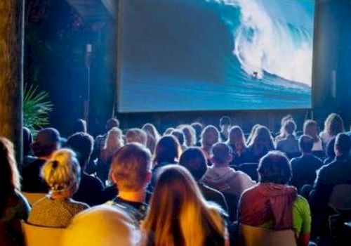 A crowd is watching a movie in an outdoor setting, featuring a large wave on the screen.