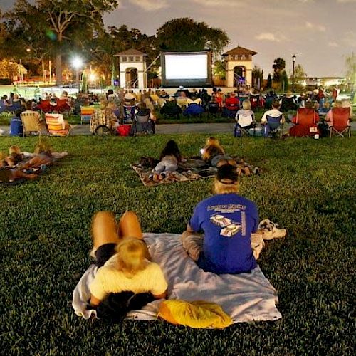 People are gathered outdoors on a lawn, sitting on chairs and blankets, enjoying a movie projected on a large screen under evening lights.