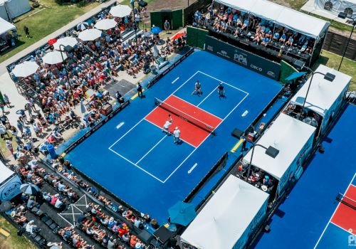 An aerial view of a pickleball court with four players competing, surrounded by a large, seated crowd and entertainment tents.