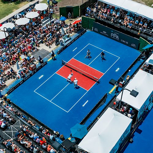 An aerial view of a pickleball court with four players competing, surrounded by a large, seated crowd and entertainment tents.