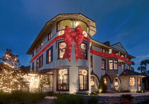 A large house decorated with a giant red ribbon and bow, illuminated by exterior lights, against a dusk sky.