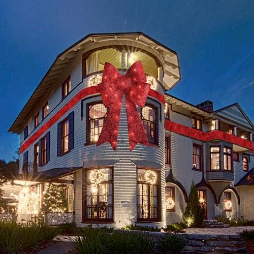 A large house decorated with a giant red ribbon and bow, illuminated by exterior lights, against a dusk sky.