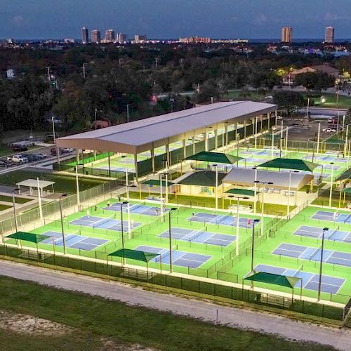 The image shows an aerial view of a large, illuminated sports complex with multiple tennis courts and some covered areas.