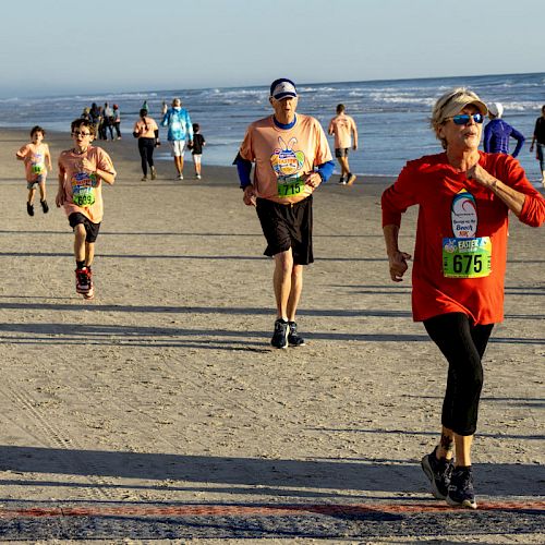People are running a race on a beach, with bib numbers visible. The ocean is on the right and more participants are in the background.