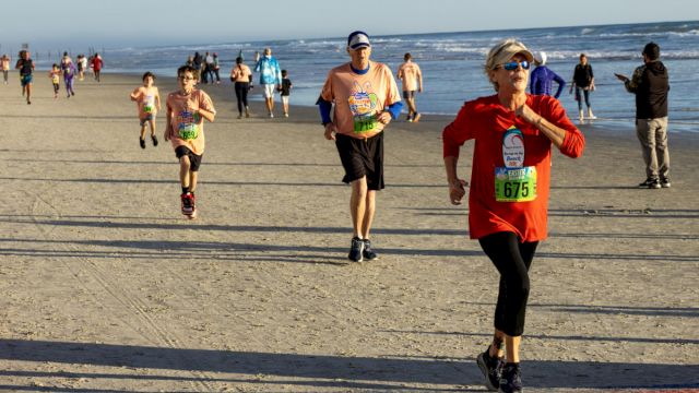 People are running a race on a beach, with bib numbers visible. The ocean is on the right and more participants are in the background.