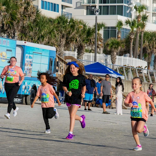 People, including children, are running a race on a beachfront. A blue truck and palm trees are visible in the background.