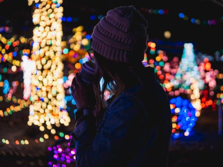 A person in a beanie enjoys a drink while illuminated by colorful festive lights in a nighttime setting.