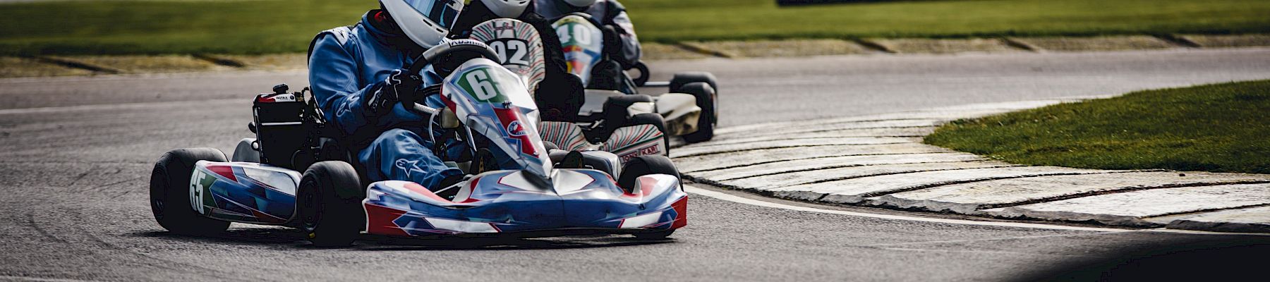 Three go-karts are racing on a track with drivers wearing helmets and racing suits, rounding a bend in overcast weather, power lines in the background.