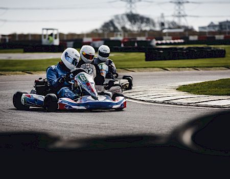 Three go-karts are racing on a track with drivers wearing helmets and racing suits, rounding a bend in overcast weather, power lines in the background.