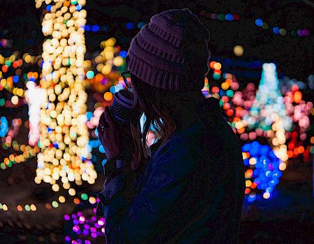 A person wearing a beanie enjoys a hot drink while gazing at colorful, festive lights in a dark environment, creating a cozy winter scene.