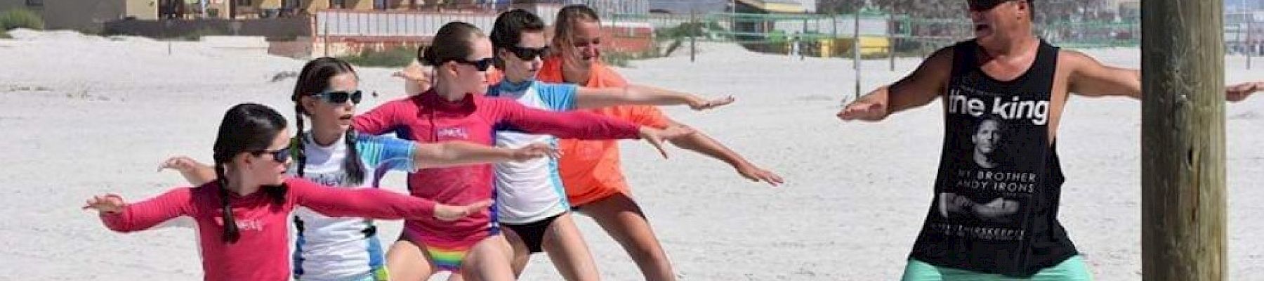 A group of kids on surfboards is participating in a beach surf lesson, following the instructions of a man standing in front of them on the sand.