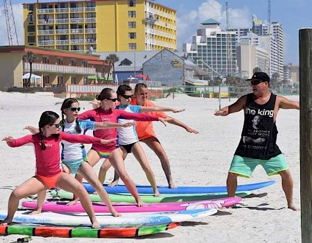 A group of kids on surfboards is participating in a beach surf lesson, following the instructions of a man standing in front of them on the sand.