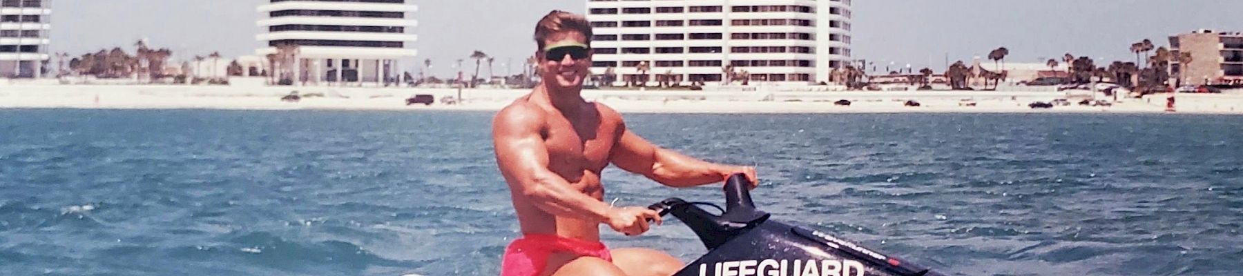 A person is riding a Yamaha lifeguard jet ski on the ocean near a coastline with high-rise buildings in the background, wearing red shorts and sunglasses.