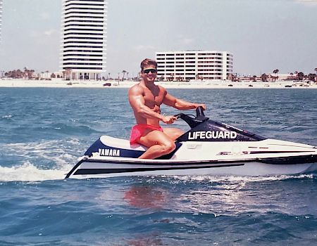 A person is riding a Yamaha lifeguard jet ski on the ocean near a coastline with high-rise buildings in the background, wearing red shorts and sunglasses.