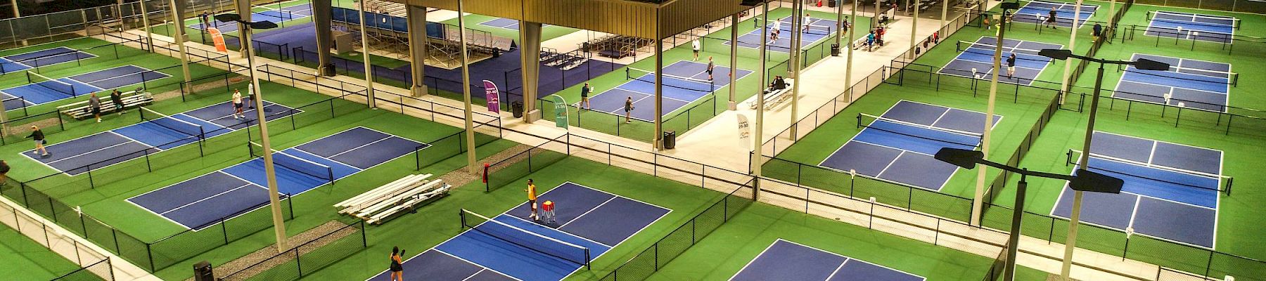 The image shows an aerial view of a well-lit pickleball facility with numerous courts, benches, and a central pavilion, taken during nighttime.