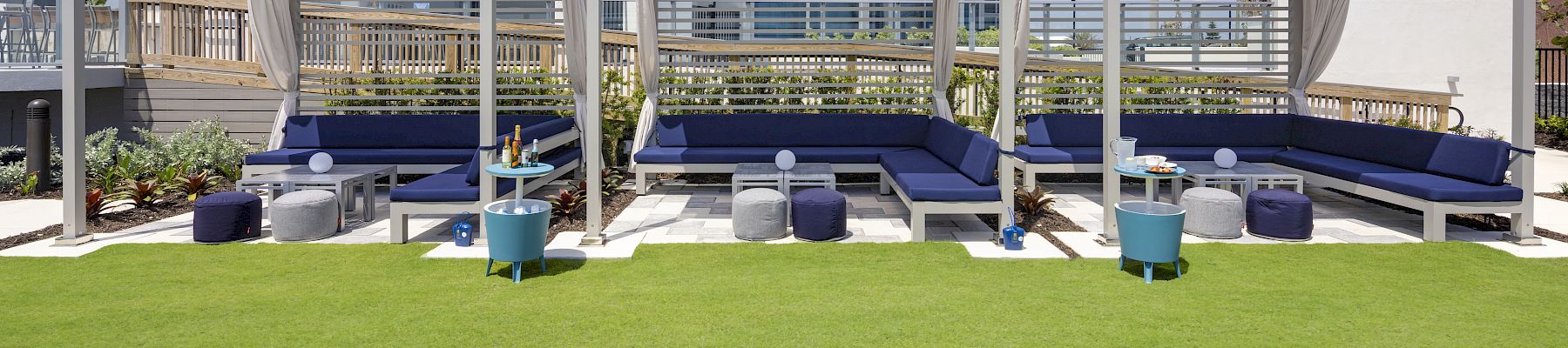 The image shows an outdoor seating area with shaded cabanas, lounge seating, small tables, and cushions, located near a modern building.