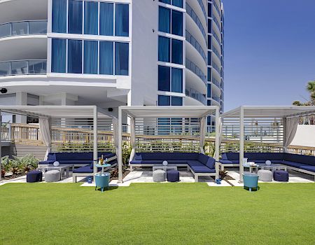 The image shows an outdoor seating area with shaded cabanas, lounge seating, small tables, and cushions, located near a modern building.