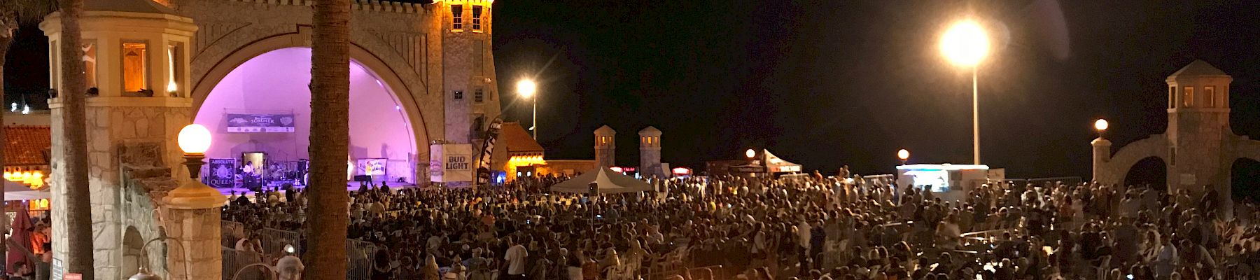 A large crowd gathers at an outdoor event in front of a lit amphitheater at night, with palm trees and illuminated street lights.