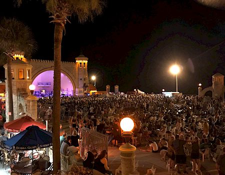 A large crowd gathers at an outdoor event in front of a lit amphitheater at night, with palm trees and illuminated street lights.