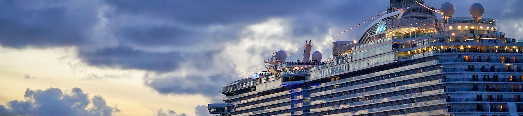 A large cruise ship sails on the ocean under a dramatic cloudy sky at sunset. The ship is well-lit and reflects on the water's surface.