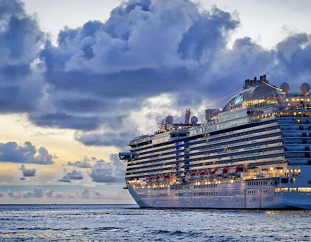 A large cruise ship sails on the ocean under a dramatic cloudy sky at sunset. The ship is well-lit and reflects on the water's surface.