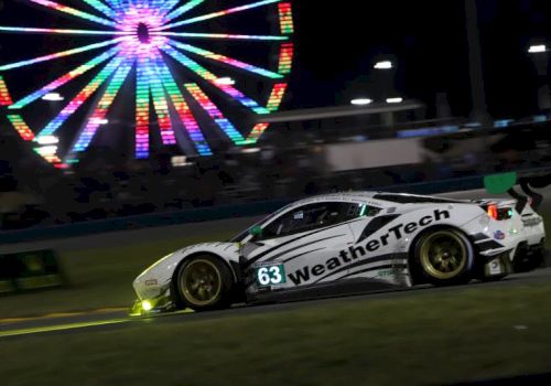 A race car speeds on a track at night while a colorful ferris-wheel light display glows in the background.
