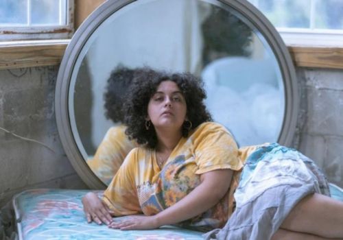 A person with curly hair is lying on a mattress in front of a round mirror, inside a room with large windows and a rustic look.
