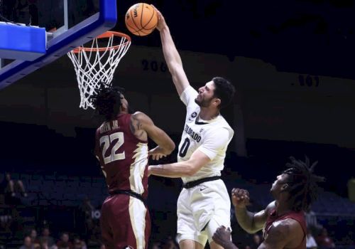 A basketball player in white jumps to dunk while a player in maroon defends. Another maroon player watches nearby.