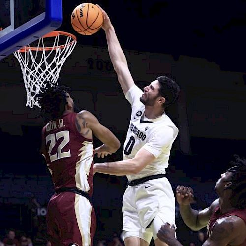 A basketball player in white jumps to dunk while a player in maroon defends. Another maroon player watches nearby.