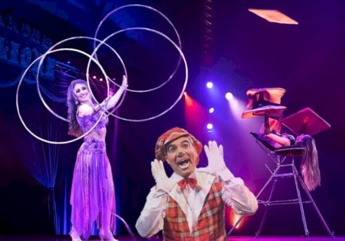 A performer is juggling hoops, another is balanced on a chair stack, and a clown poses in front of a brightly lit stage backdrop.