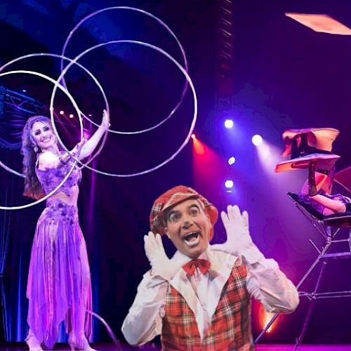 A performer is juggling hoops, another is balanced on a chair stack, and a clown poses in front of a brightly lit stage backdrop.